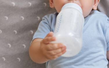 Upper view of adorable baby boy holding bottle with formula lying in crib, enjoying milk or tasty healthy nutritious baby formula. Bottle-feeding concept. Childcare