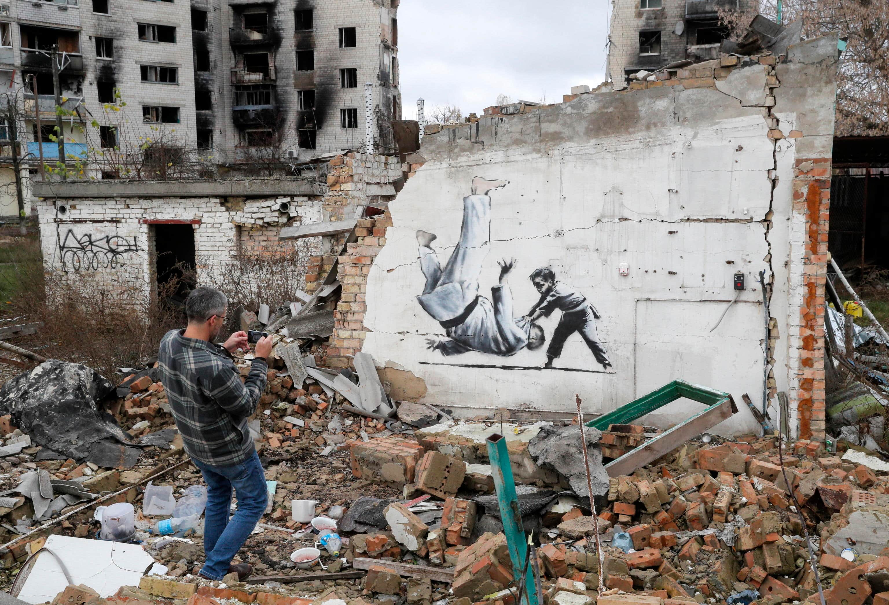 epa10303647 A man takes a picture of a mural depicting child overthrowing a man in judo clothes, in a style resembling the works of British street artist Banksy, at a wall of a building that was destroyed during shelling in Borodyanka, Ukraine, 13 November 2022. A photo of a mural in Borodyanka depicting a gymnast was shared by Banksy on his social media channel on 11 November, while other works attributed to him in different locations in Ukraine and resembling Banksy's style have not been shared on the artist's channel yet. Russian troops entered Ukraine on 24 February 2022, starting a conflict that has provoked destruction and a humanitarian crisis.  EPA/SERGEY DOLZHENKO