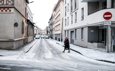 Wide view of a snow covered urban street with a pedestrian crossing an icy road during a weather alert in Clermont Ferrand, Puy de Dome, France on January 6, 2026. The scene shows slippery conditions with snow and ice on the roadway highlighting risks for pedestrians and vehicles during a snow and ice warning. (Photo by Romain Costaseca / Hans Lucas / AFP via Getty Images)